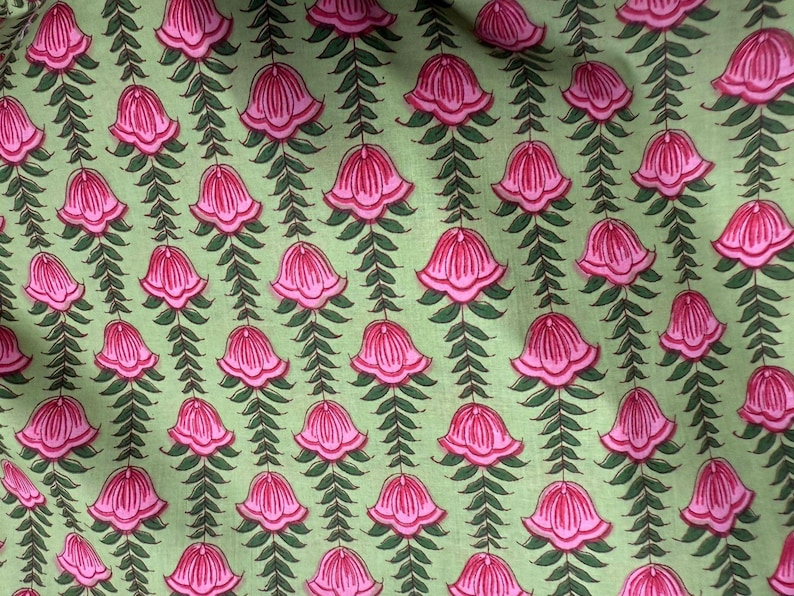 May include: Close-up of a fabric with a repeating floral pattern. The design features pink bell-shaped flowers with green leaves and stems against a light green background. The pattern is symmetrical and detailed.