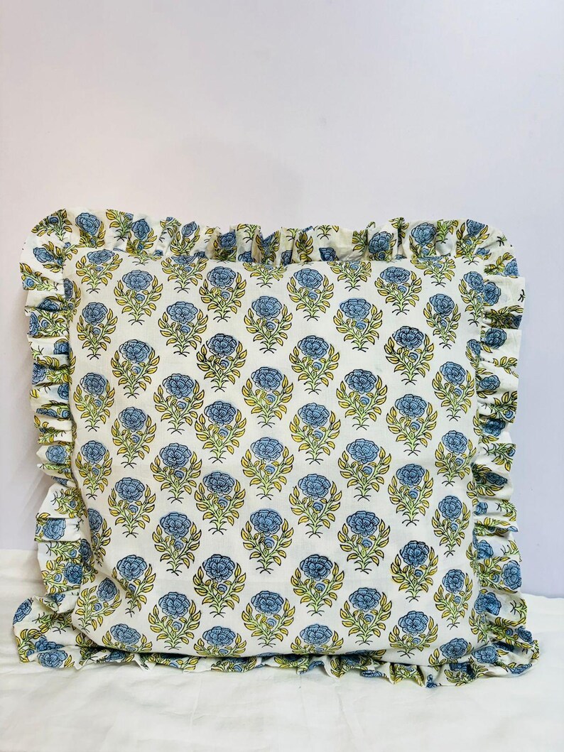 May include: A square throw pillow featuring a repeating floral pattern of blue blossoms and green leaves on a white background. The pillow is finished with a gathered fabric ruffle trim along all four edges, matching the floral print of the cover.