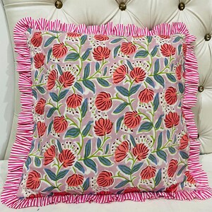 May include: A square throw pillow featuring a light purple background with a repeating pattern of coral-colored flowers and green leaves. The pillow is bordered by a ruffled edge made of fabric with thin, alternating white and pink vertical stripes.