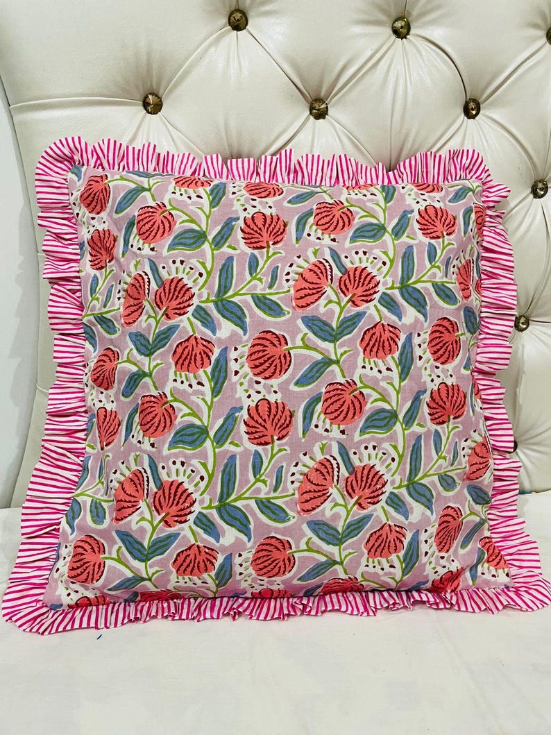 May include: A square throw pillow featuring a light purple background with a repeating pattern of coral-colored flowers and green leaves. The pillow is bordered by a ruffled edge made of fabric with thin, alternating white and pink vertical stripes.