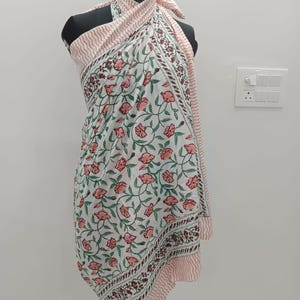 May include: A white sarong featuring a repeating floral pattern with pink flowers and green vines. The fabric has a border with a pink and white wavy line design. The sarong is draped around a mannequin, tied at the neck with a halter-style knot.
