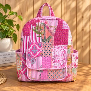 Puede incluir: Una mochila acolchada con un diseño de patchwork vibrante en varios tonos de rosa, blanco y naranja. La bolsa incluye un asa superior, dos bolsillos laterales y un bolsillo frontal con solapa, todo construido con costuras verticales.