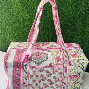 May include: A rectangular quilted duffel bag featuring a white base with pink and green floral block print patterns. The bag includes two long handles with a pink and white striped design, a zippered top closure, and matching striped trim along the edges.