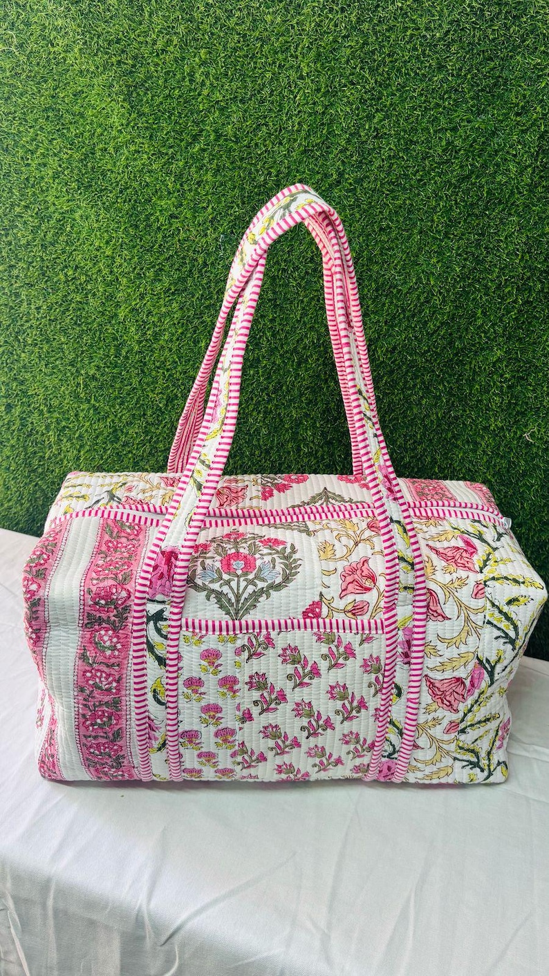 May include: A rectangular quilted duffel bag featuring a white base with pink and green floral block print patterns. The bag includes two long handles with a pink and white striped design, a zippered top closure, and matching striped trim along the edges.