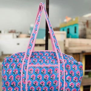 May include: A blue duffel bag with a pink floral pattern. The bag has pink handles and trim. The floral pattern is repeated across the bag's surface. The bag is made of a quilted fabric.