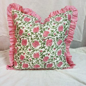 May include: A square throw pillow featuring a cream-colored base with a repeating pattern of pink tulips and green vines. The pillow is bordered by a ruffled edge made of fabric with thin, vertical red and white stripes on all four sides.