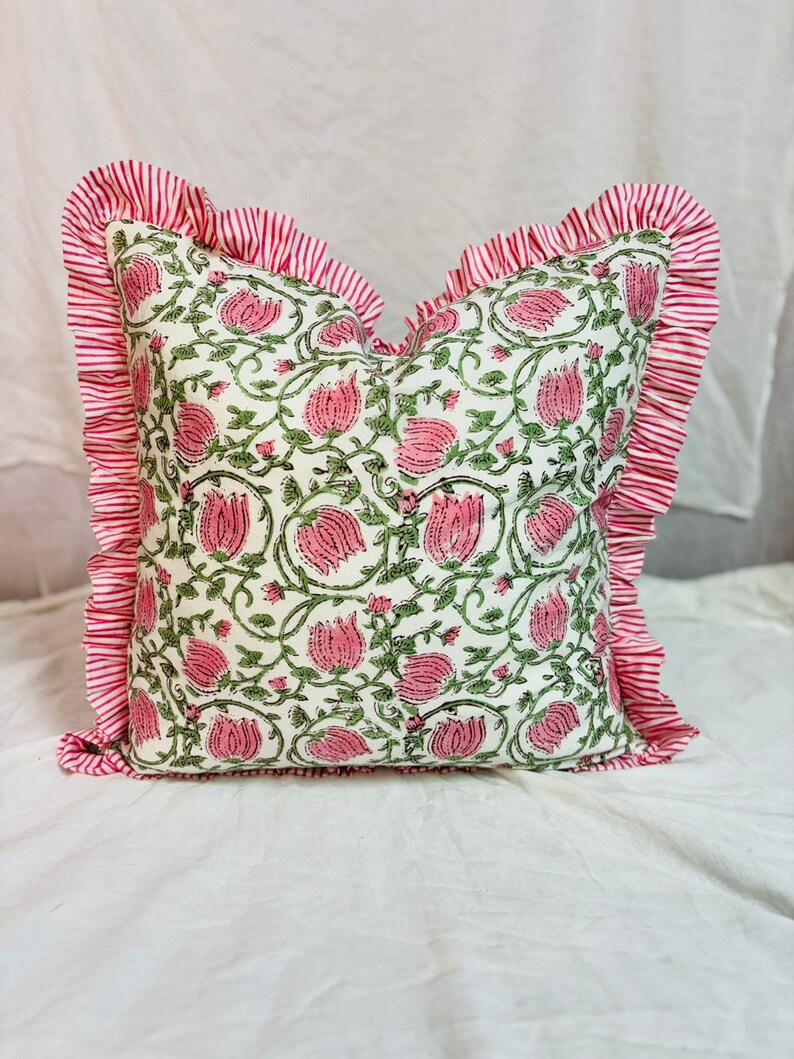 May include: A square throw pillow featuring a cream-colored base with a repeating pattern of pink tulips and green vines. The pillow is bordered by a ruffled edge made of fabric with thin, vertical red and white stripes on all four sides.