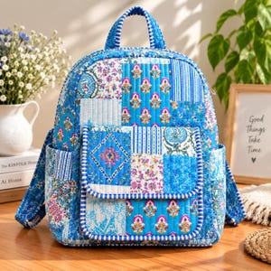 Puede incluir: Una mochila acolchada con un diseño de patchwork en tonos azules, blancos y rosas. La bolsa cuenta con un asa superior, correas ajustables para los hombros, dos bolsillos laterales y un bolsillo frontal con solapa, decorados con varios patrones florales.