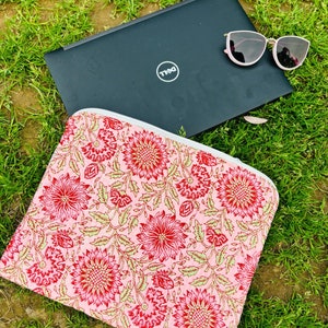 Könnte beinhalten: Eine rosa Laptop-Hülle mit Blumenmuster und Reißverschluss. Die Hülle liegt auf einem Bett aus grünem Gras mit einem schwarzen Laptop und einer rosa Sonnenbrille darauf. Der Laptop hat ein Dell-Logo auf dem Deckel.