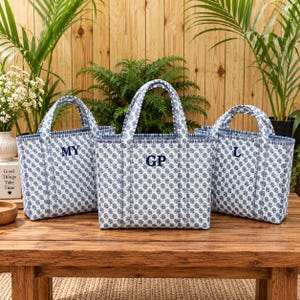 Puede incluir: Tres bolsos tote acolchados con un estampado floral azul y blanco. Cada bolso tiene dos asas y está bordado con diferentes iniciales: MY, GP y L. Los bolsos están estructurados con una base rectangular y detalles de costuras verticales.