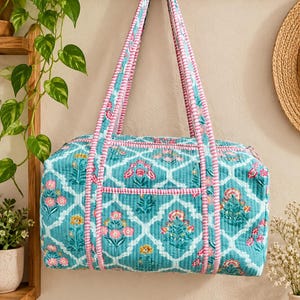 May include: A quilted duffel bag featuring a teal background with a white lattice pattern and floral motifs in pink, yellow, and green. The bag includes two long shoulder straps with a pink and white striped trim that matches the piping.
