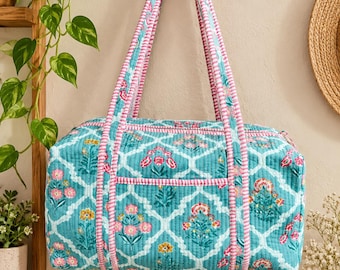 May include: A quilted duffel bag featuring a teal background with a white lattice pattern and floral motifs in pink, yellow, and green. The bag includes two long shoulder straps with a pink and white striped trim that matches the piping.