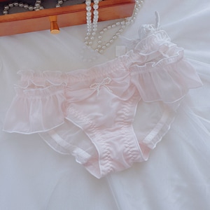 Puede incluir: Ropa interior de encaje rosa con un lazo y un adorno de perlas. La ropa interior tiene un borde con volantes y una tela transparente.