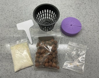 May include: A kit for growing plants includes a black plastic net pot, a purple foam disc, a small bag of white fertilizer, a small bag of brown clay pebbles, a small bag of seeds, and a white plastic plant label.