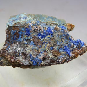 May include: A close-up of a mineral specimen featuring a mix of colors. The rock has a rough, textured surface with areas of deep blue, turquoise, brown, and white. The blue crystals are clustered throughout the stone.
