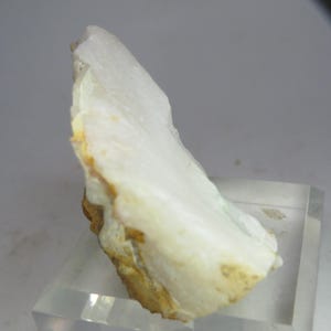 Op de afbeelding: Een wit, onregelmatig gevormd mineraal specimen met een gelaagde textuur. De steen heeft gebieden met lichtbruine en gele tinten langs de randen. Het mineraal wordt tentoongesteld op een heldere, rechthoekige standaard.