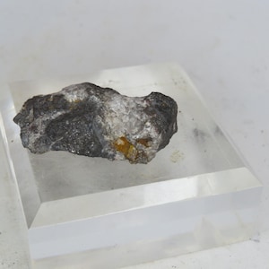 Può includere: Una roccia grezza di forma irregolare, con una miscela di colori grigio scuro, argento e bianco, con sfumature di giallo e marrone. La roccia è esposta su un blocco acrilico trasparente e rettangolare, che ne mette in risalto la consistenza naturale.