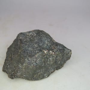 Puede incluir: Una roca de color gris oscuro, de forma irregular y superficie texturizada. La roca tiene pequeñas motas de gris claro y blanco. El fondo es blanco liso.