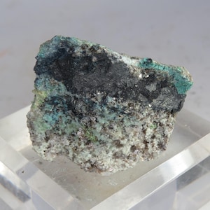 May include: A rough-hewn mineral specimen with a dark, almost black, center and a mix of teal and light green hues around the edges. The base of the rock is a speckled, light gray color. The mineral sits on a clear, transparent surface.