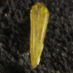 May include: A translucent, yellow crystal with a pointed top and a faceted body. The crystal is elongated and tapers towards the bottom, resting on a dark, textured surface. The crystal's color is a vibrant, golden yellow.