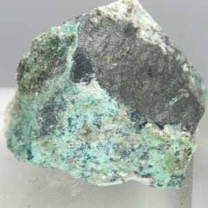 May include: A close-up of a mineral specimen with a variety of colours and textures. The rock has a dark grey, almost black, central area, surrounded by patches of vibrant turquoise and hints of white. The surface is rough and uneven, showcasing the natural formation.