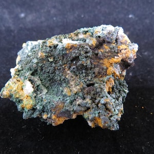 Puede incluir: Primer plano de un espécimen mineral con una superficie rugosa y texturizada. La roca presenta una mezcla de colores, incluyendo verde oscuro, naranja y blanco, con un aspecto moteado. El mineral parece ser una formación natural.