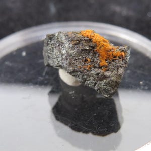 Può includere: Un piccolo sasso grigio scuro con una macchia di cristalli arancioni brillanti. Il sasso è appoggiato su una superficie bianca.