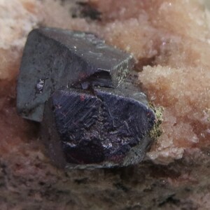 Peut inclure: Un cristal minéral de couleur gris foncé et métallique, de forme géométrique pointue. Le cristal est incrusté dans une matrice de couleur rose clair et de texture rugueuse.