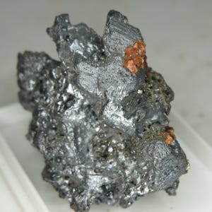 Puede incluir: Primer plano de un espécimen mineral con un brillo metálico gris oscuro. La formación rocosa tiene una superficie rugosa y texturizada con pequeñas inclusiones de color marrón rojizo. La forma general es irregular y dentada, lo que sugiere un estado natural y sin pulir.