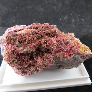 May include: A close-up of a mineral specimen featuring a mix of colors. The rock has a base of gray and brown, with vibrant red and pink crystalline formations. Small white and yellow patches are also visible.