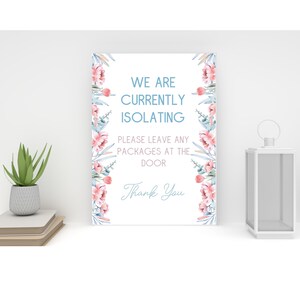 May include: A white sign with the text "WE ARE CURRENTLY ISOLATING, PLEASE LEAVE ANY PACKAGES AT THE DOOR, Thank You." The sign is decorated with pink and blue floral accents. A small potted plant and a white lantern are also visible.