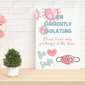 May include: Pink and blue sign with the text "I am currently isolating. Please leave any packages at the door. Be safe."  The sign also features a pink heart, a pink leopard print face mask, and pink and blue virus illustrations.