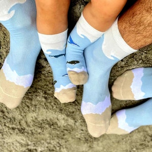 Puede incluir: Un conjunto de cuatro calcetines azules con un diseño de playa. Los calcetines presentan nubes blancas, arena marrón y ballenas azules.