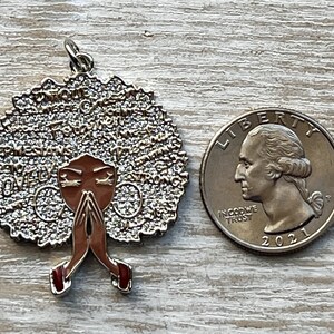 May include: A gold-toned pendant featuring a woman with brown skin and a large afro. The afro is covered in small, sparkling rhinestones. The woman is wearing a red dress and has her hands clasped in prayer. The pendant is about the size of a quarter.