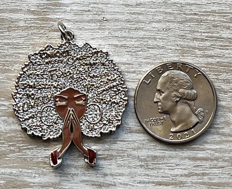 May include: A gold-toned pendant featuring a woman with brown skin and a large afro. The afro is covered in small, sparkling rhinestones. The woman is wearing a red dress and has her hands clasped in prayer. The pendant is about the size of a quarter.