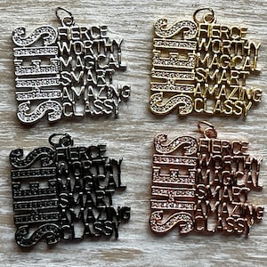 May include: Four charm pendants with the word "SHE" in large letters and the words "Fierce", "Worthy", "Magical", "Smart", "Amazing", and "Classy" in smaller letters. The charms are in silver, gold, black, and rose gold.