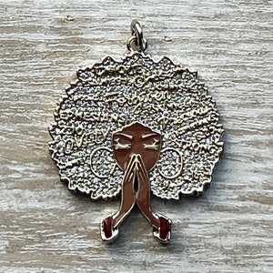 May include: Gold-tone pendant featuring a stylized image of a woman with an afro hairstyle. The afro is covered in small, sparkling rhinestones and the woman's face is brown with closed eyes. The woman is depicted with her hands clasped in prayer. The pendant has the words "QUEEN" and "STRONG" written around the afro.