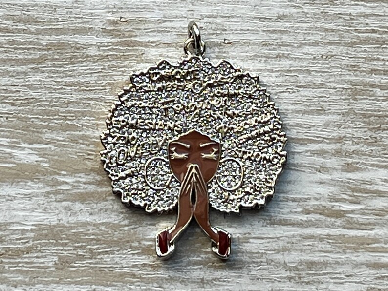 May include: Gold-tone pendant featuring a stylized image of a woman with an afro hairstyle. The afro is covered in small, sparkling rhinestones and the woman's face is brown with closed eyes. The woman is depicted with her hands clasped in prayer. The pendant has the words "QUEEN" and "STRONG" written around the afro.