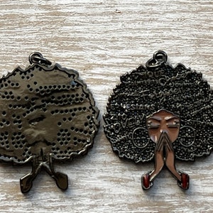 May include: Black metal charm with a woman's face and afro hair. The hair is covered in words like "strong", "love", and "queen". The back of the charm is plain.
