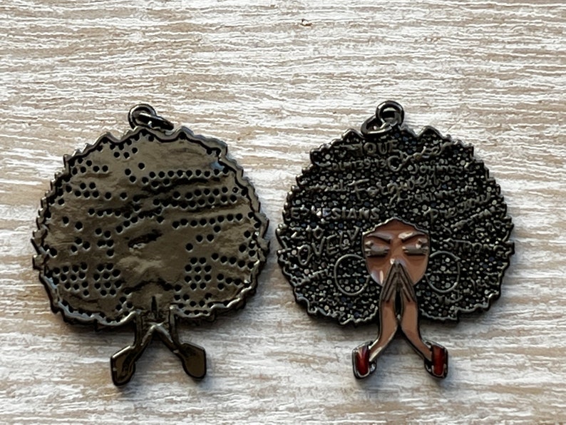May include: Black metal charm with a woman's face and afro hair. The hair is covered in words like "strong", "love", and "queen". The back of the charm is plain.