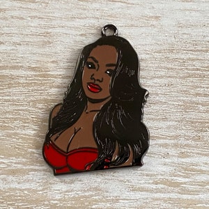 May include: A black metal charm featuring a portrait of a woman with long black hair, wearing a red strapless top.