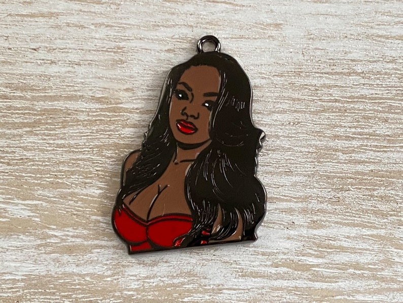 May include: A black metal charm featuring a portrait of a woman with long black hair, wearing a red strapless top.