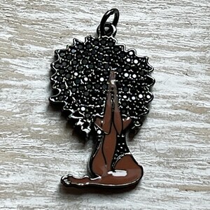 Pode incluir: Um amuleto de metal preto que representa uma mulher em uma pose de ioga com as m&atilde;os juntas em ora&ccedil;&atilde;o. Seu cabelo est&aacute; penteado em um afro e &eacute; representado por um design preto brilhante.