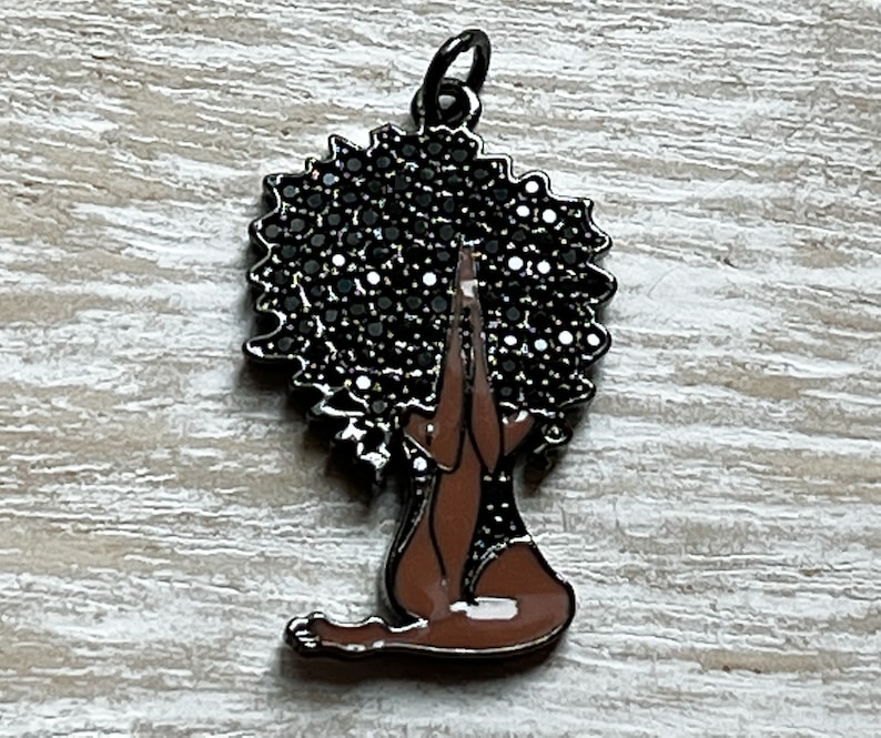 Pode incluir: Um amuleto de metal preto que representa uma mulher em uma pose de ioga com as m&atilde;os juntas em ora&ccedil;&atilde;o. Seu cabelo est&aacute; penteado em um afro e &eacute; representado por um design preto brilhante.