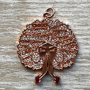May include: A gold-tone pendant featuring a stylized image of a woman with an afro hairstyle. The afro is covered in small, clear rhinestones and the woman's face is partially obscured by her hands, which are clasped in prayer. The pendant has a loop at the top for attaching to a chain.
