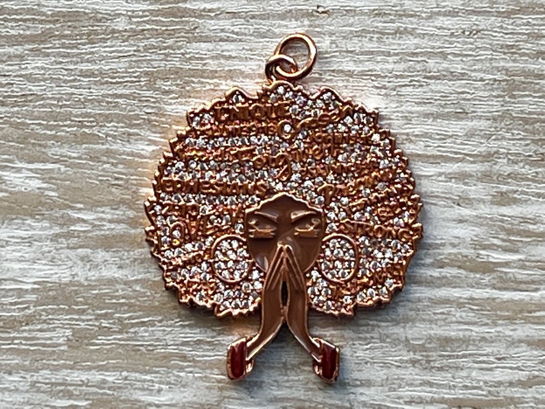 May include: A gold-tone pendant featuring a stylized image of a woman with an afro hairstyle. The afro is covered in small, clear rhinestones and the woman's face is partially obscured by her hands, which are clasped in prayer. The pendant has a loop at the top for attaching to a chain.