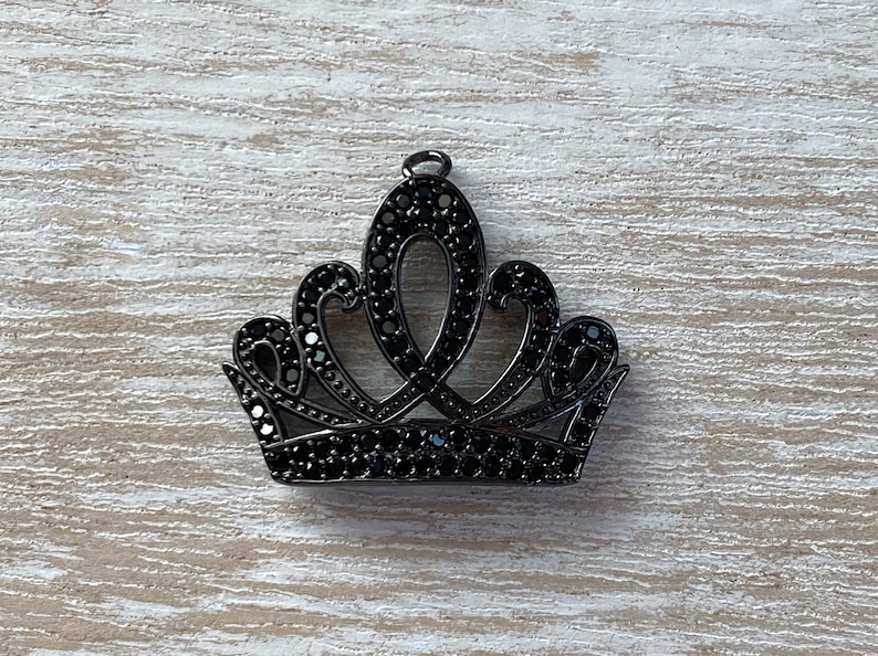 May include: A black metal crown charm with black rhinestone accents. The crown has a pointed top and a rounded base.