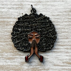 May include: A black metal charm with a woman's face and afro hair. The hair is covered in words like "love", "strong", "beautiful", and "queen". The woman is praying with her hands clasped together. The charm has a gold finish.