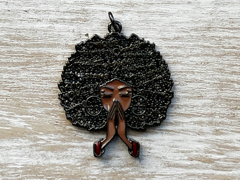 May include: A black metal charm with a woman's face and afro hair. The hair is covered in words like "love", "strong", "beautiful", and "queen". The woman is praying with her hands clasped together. The charm has a gold finish.