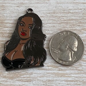 May include: A black metal charm featuring a portrait of a woman with long black hair, wearing a black dress and red lipstick.